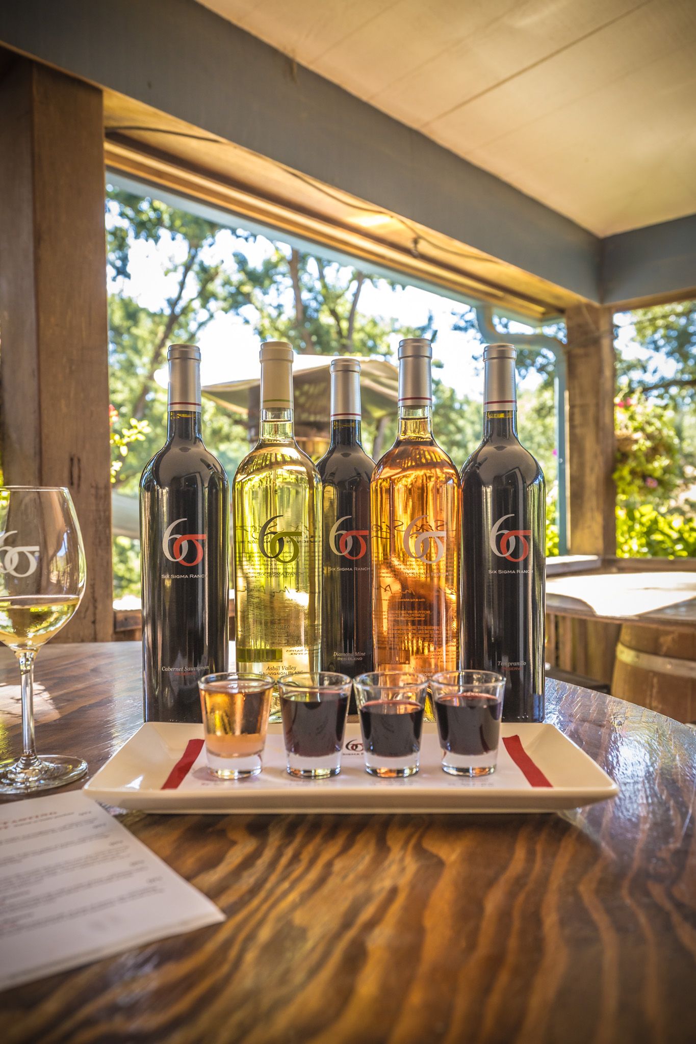 Wine bottles and tasting samples arranged on a wooden table with sunlit windows in background.