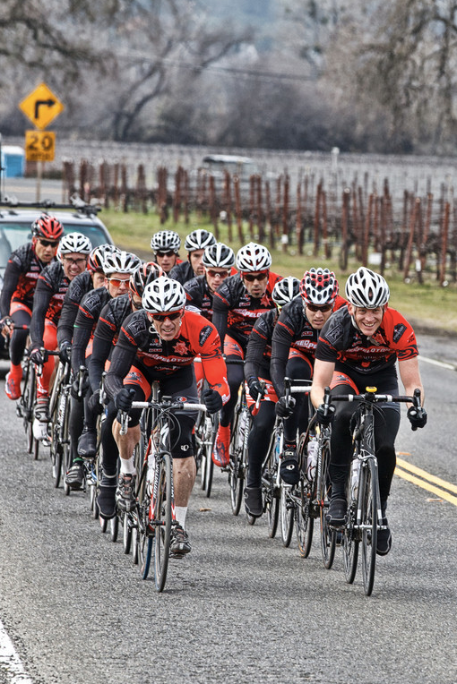 Group of cyclists in red jerseys riding in tight formation on a wet road near a vineyard.