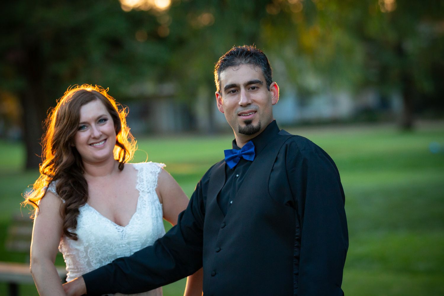A couple stands together in formal wedding attire against a green outdoor backdrop.