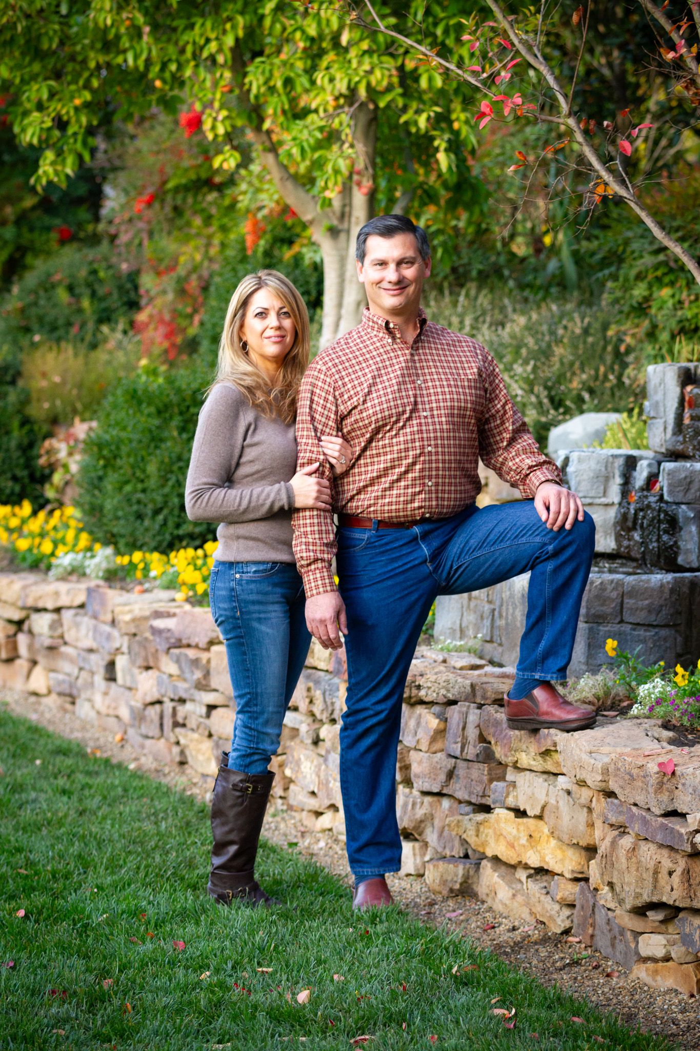 Casual portrait of couple posing by stone retaining wall with yellow flowers and trees in background.