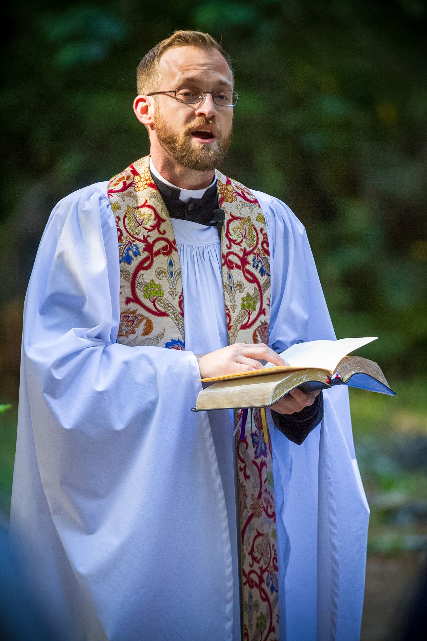 Officiant in white robe and decorative stole reads from book during outdoor ceremony.
