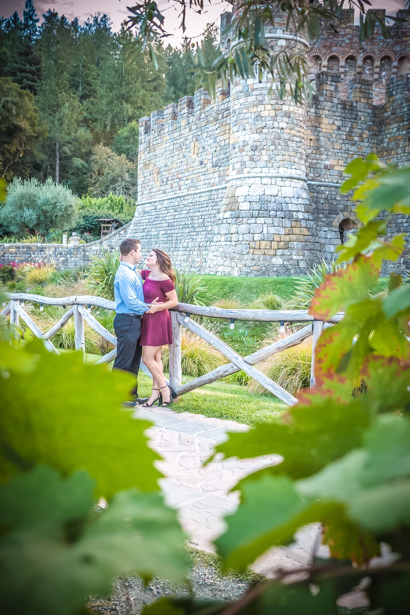 Couple poses romantically on bridge near stone castle wall surrounded by lush greenery.
