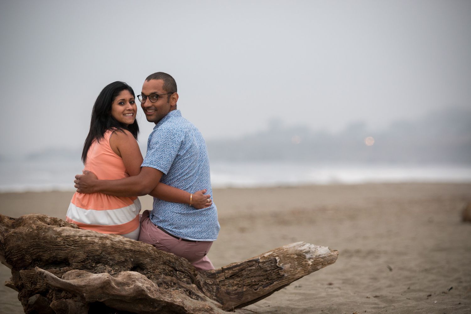 A couple sitting together on driftwood at the beach.