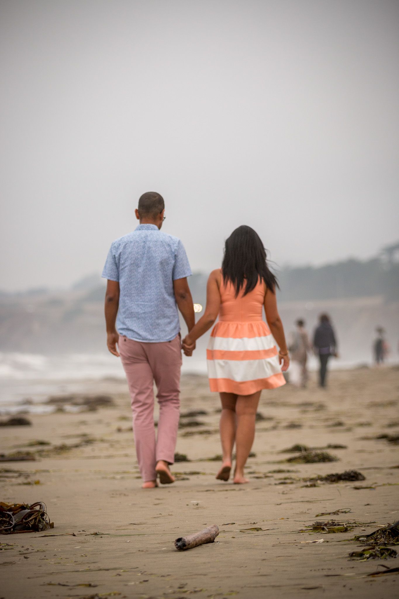 A couple walking hand in hand on a misty beach wearing casual clothing.