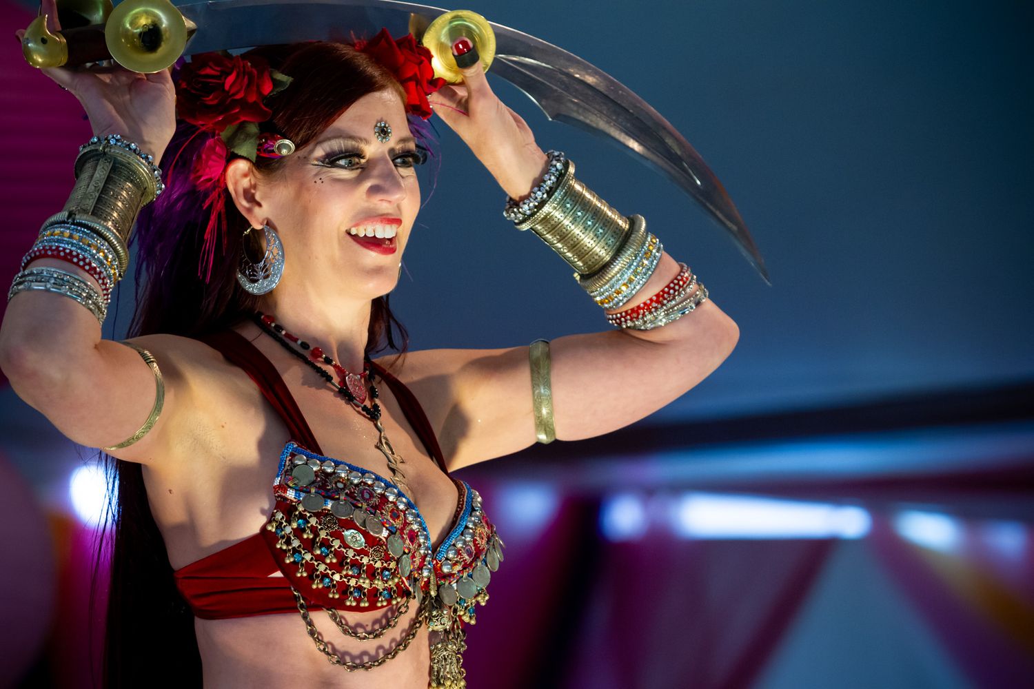 A belly dancer performs in costume with golden props raised above her head against a dark blue background.