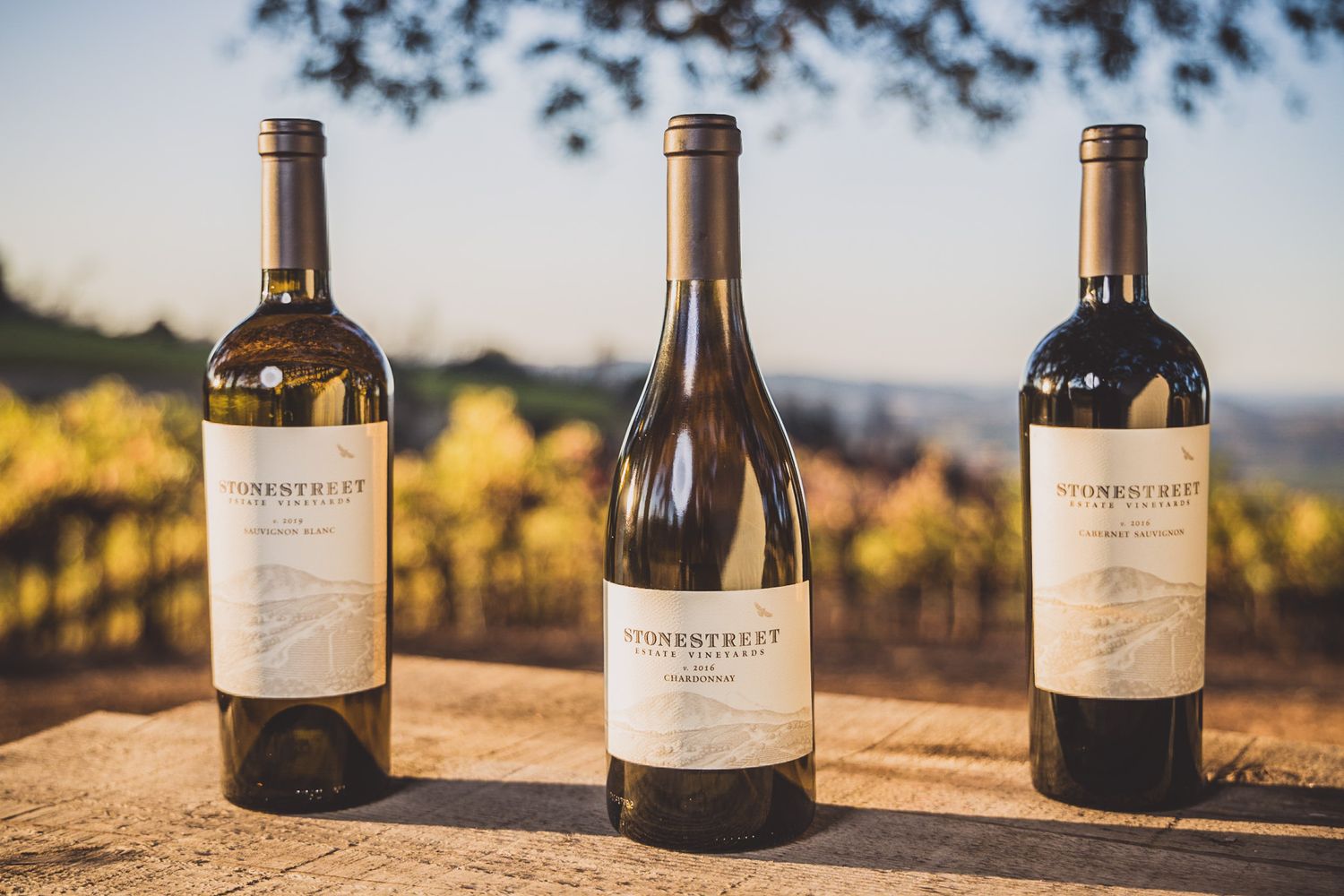 Three wine bottles displayed on outdoor table overlooking vineyard landscape.
