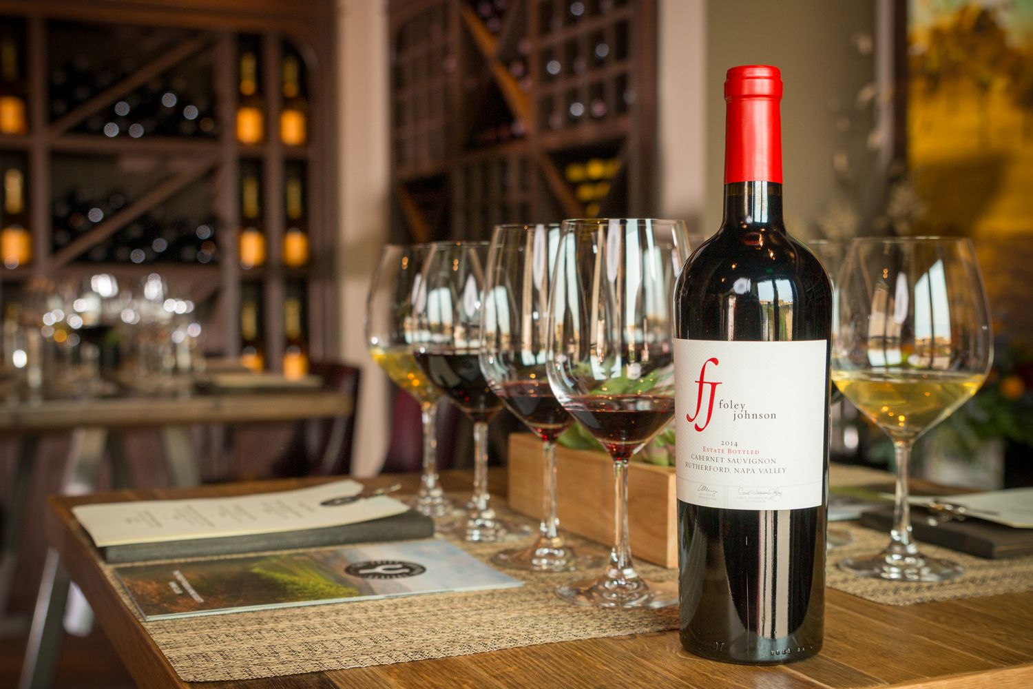Wine bottle and glasses arranged for tasting on wooden table with wine racks in background.