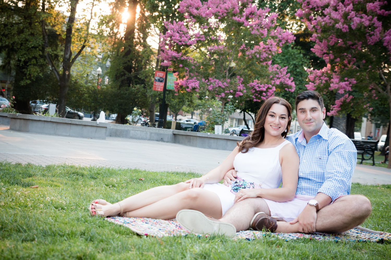 Couple sitting on blanket in park at sunset with pink flowering trees and soft sunlight in background.