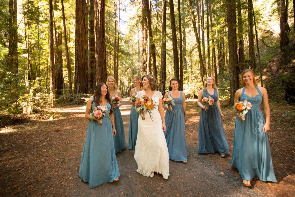 Wedding party walks through sunlit forest carrying colorful bouquets of orange and pink flowers.