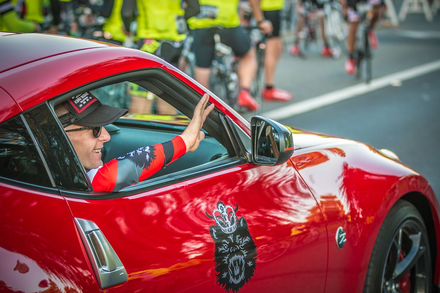 A red sports car driver participates in a cycling event with cyclists visible in background.