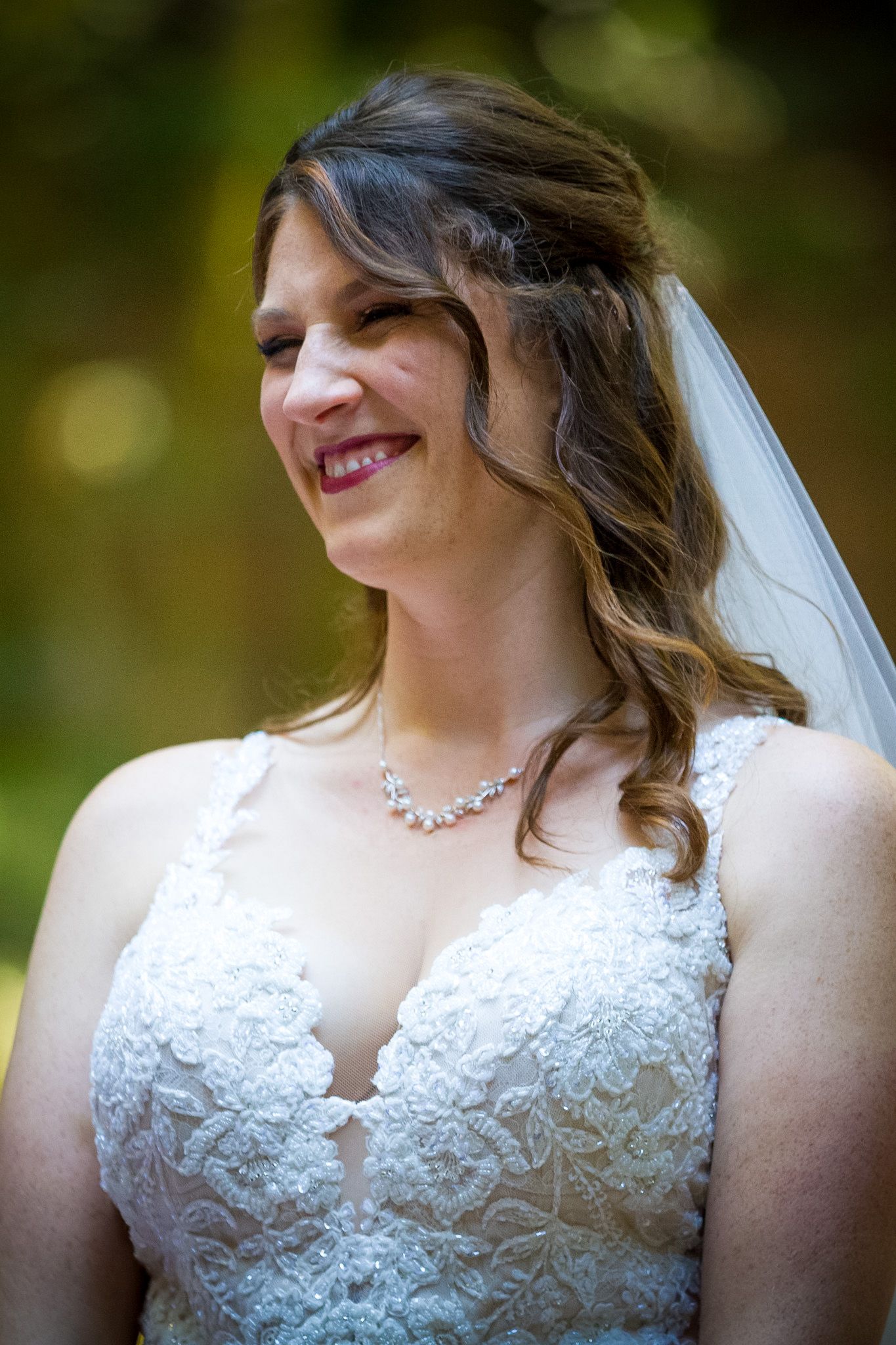 Bride wearing lace wedding gown and veil smiles in outdoor forest setting.