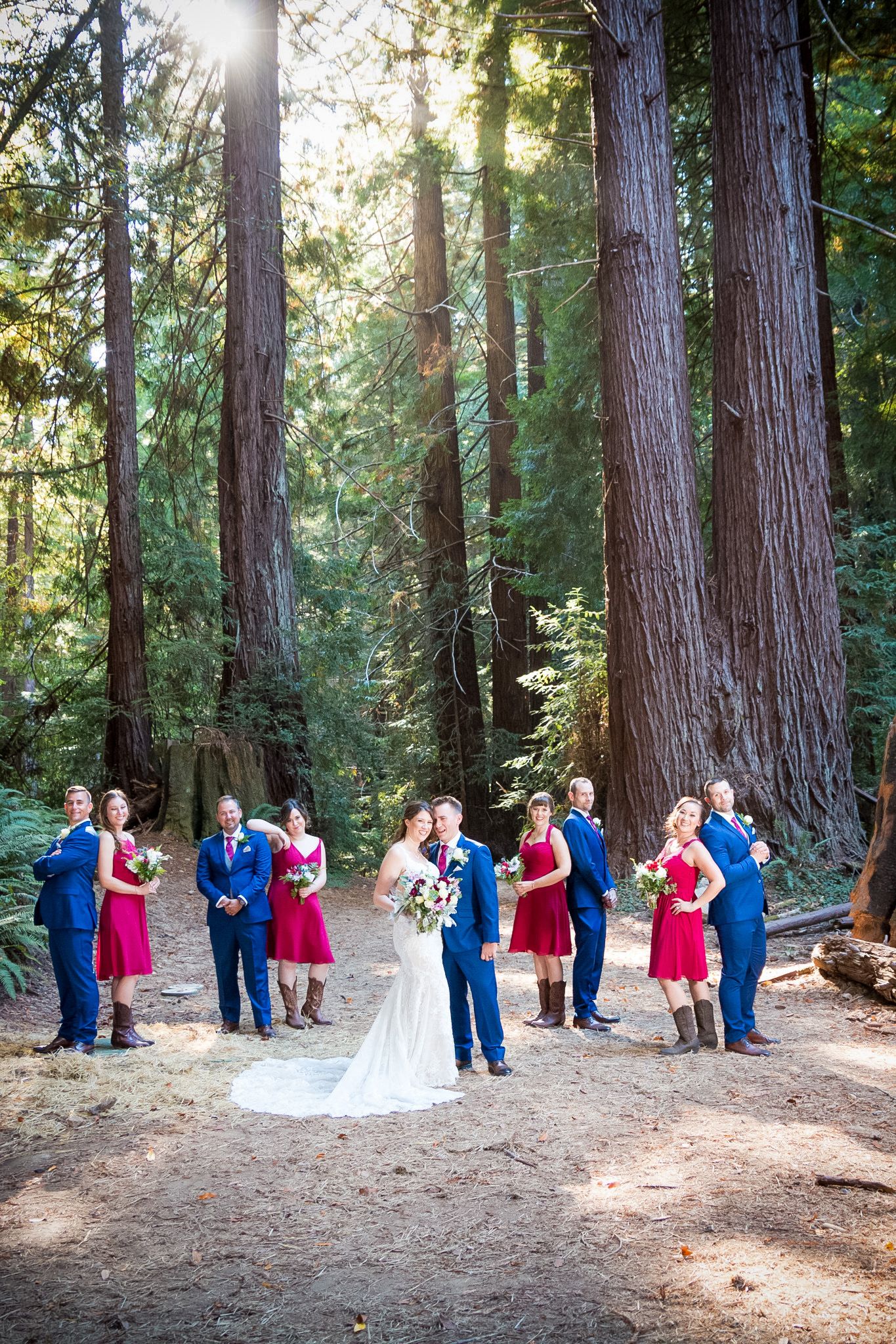 Sunlit forest wedding photo with bridal party in red dresses and navy suits among tall redwoods.