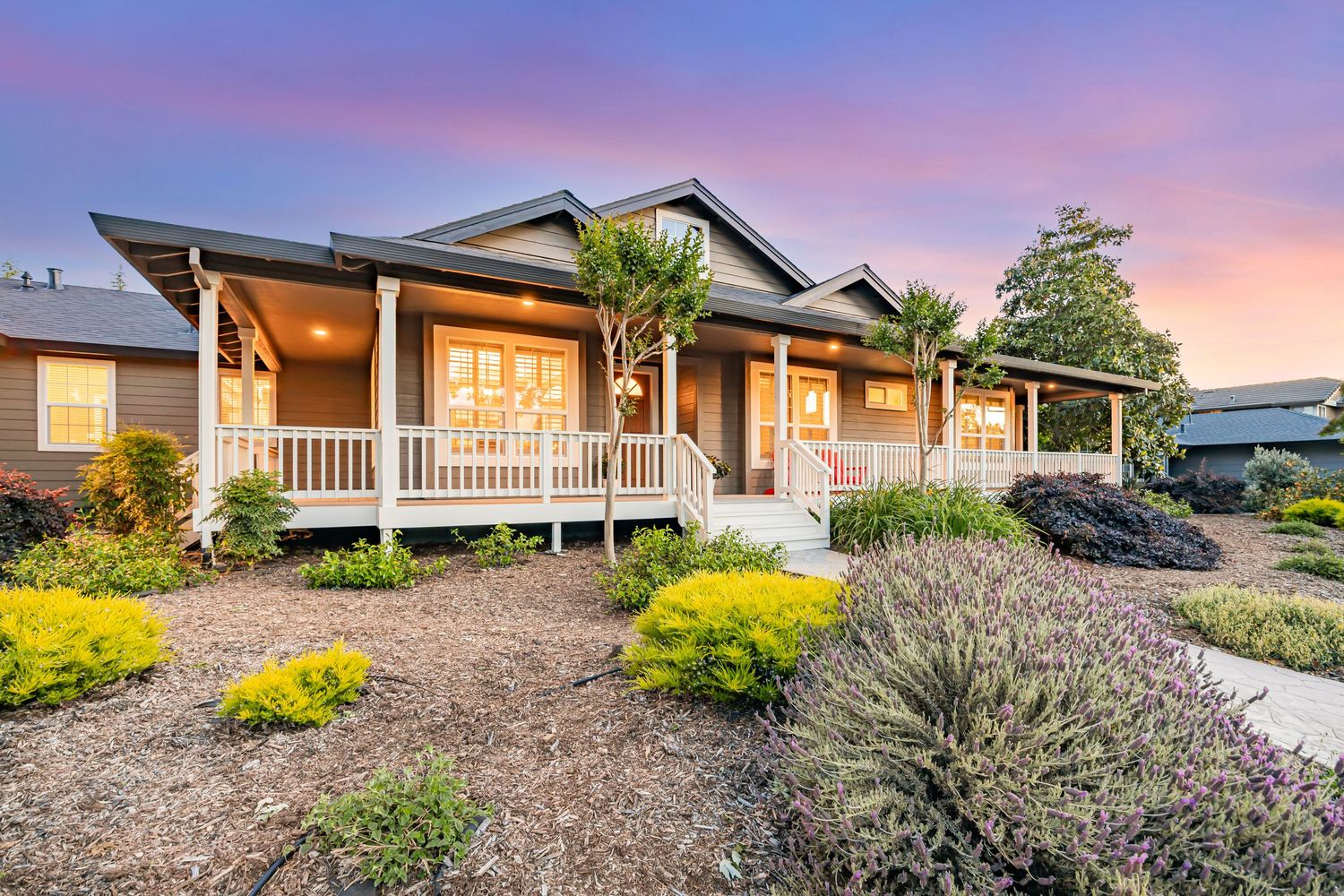 Charming ranch house with wraparound porch and drought-tolerant garden under pink sky.