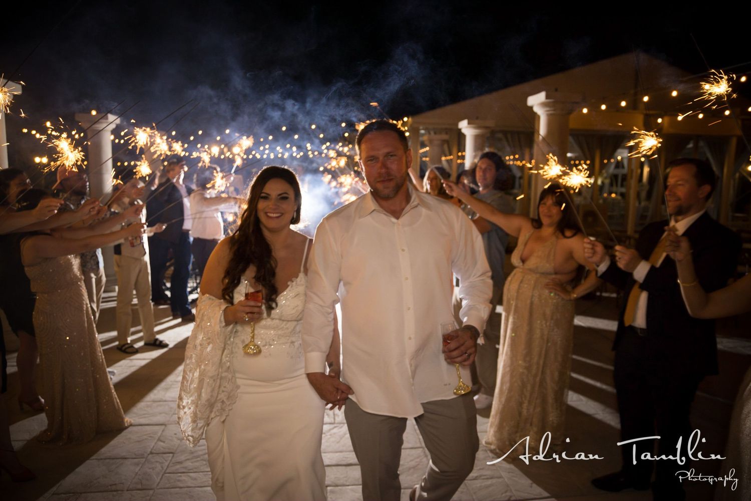 Newlyweds celebrate during their sparkler exit under twinkling lights at dusk.