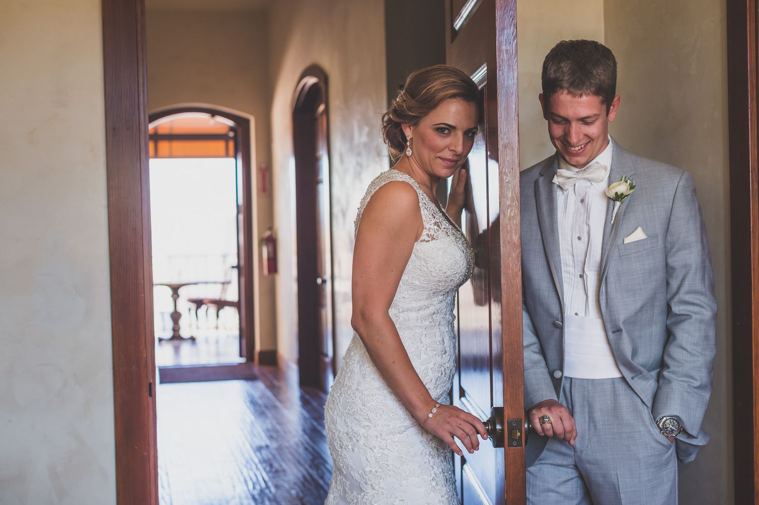 Bride and groom share a private moment against a white wall in a hallway, dressed in white and light gray attire.