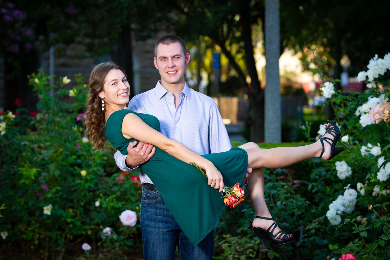 Man carrying woman in green dress playfully among blooming roses in garden setting.
