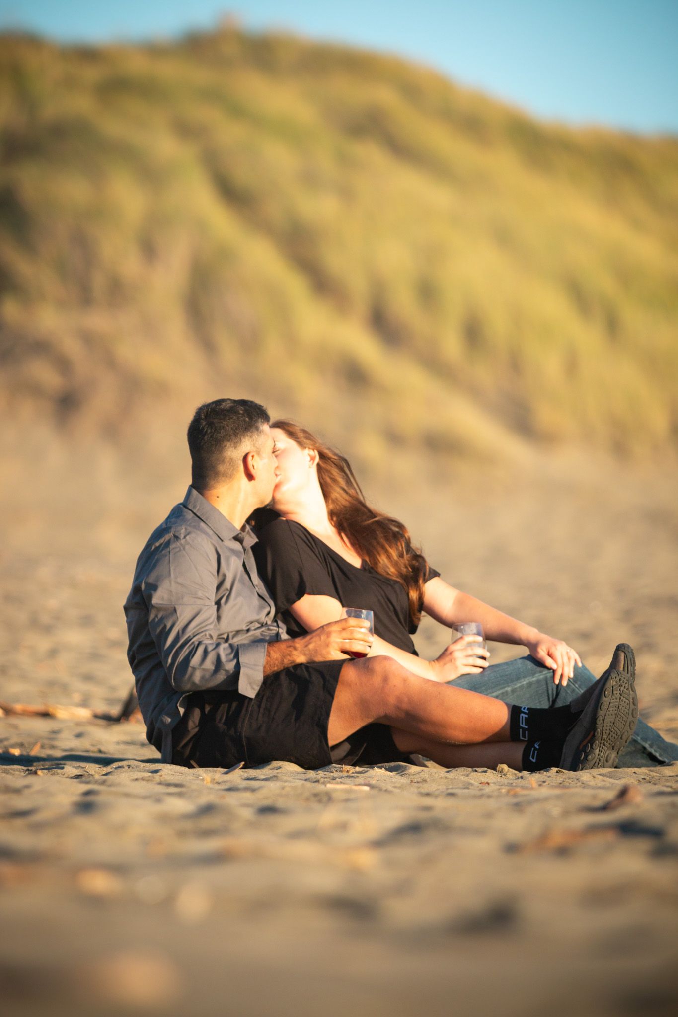 Couple sitting close together on sandy beach with coastal cliffs in background.