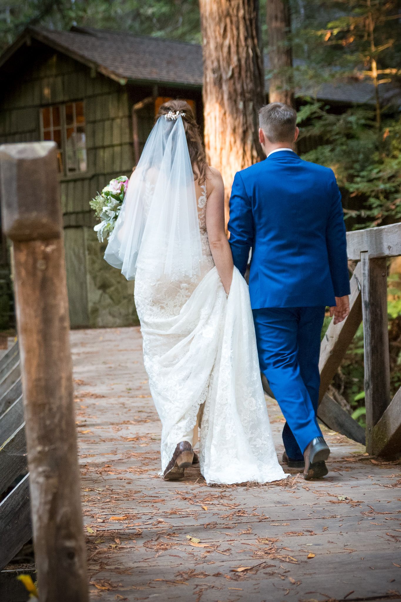 A couple walks along a wooden boardwalk near a rustic cabin, with the bride in white and groom in blue.