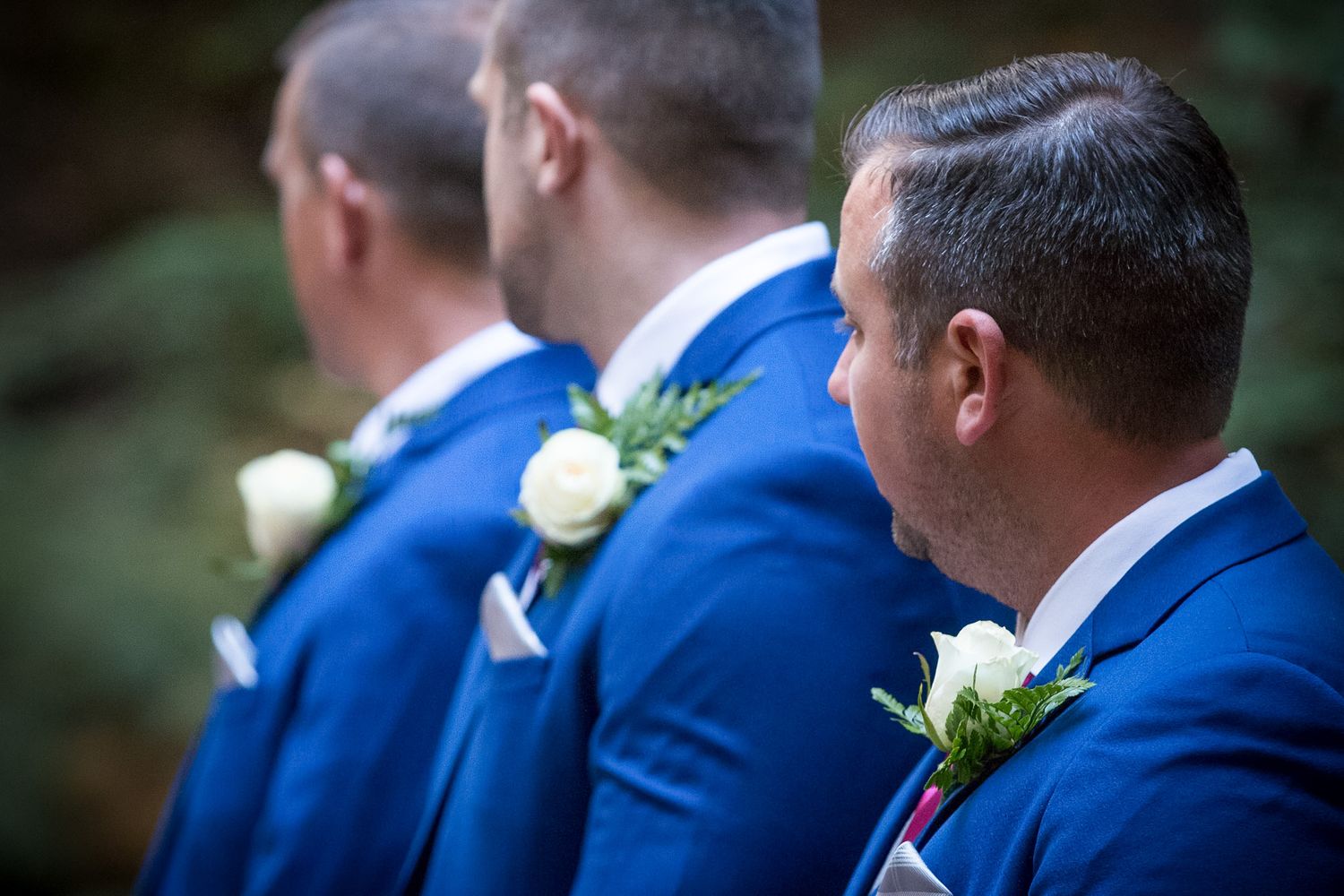 Groomsmen wearing matching blue suits with white rose boutonnieres at outdoor wedding.