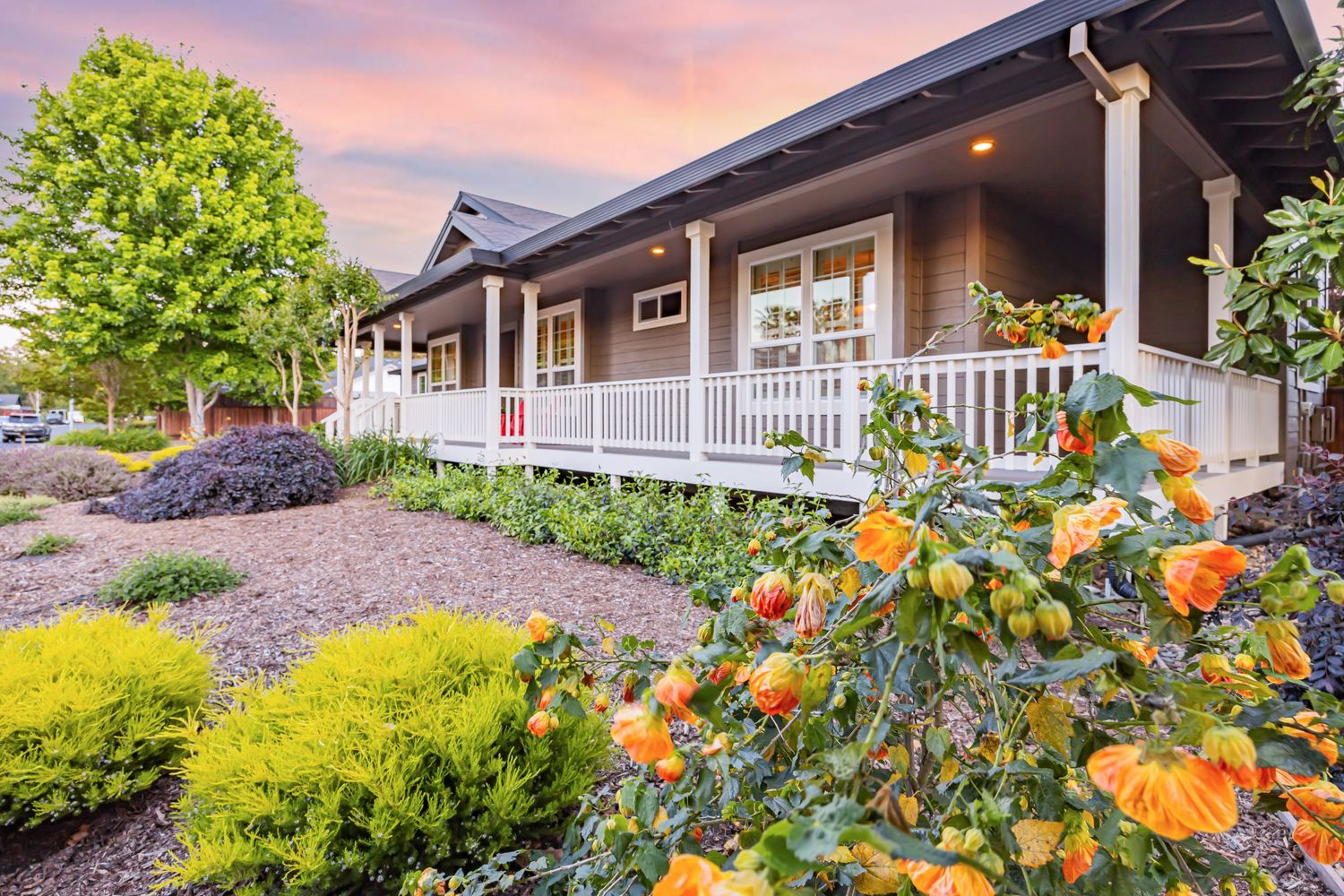 Charming ranch house with white wraparound porch and colorful garden featuring orange flowers at sunset.