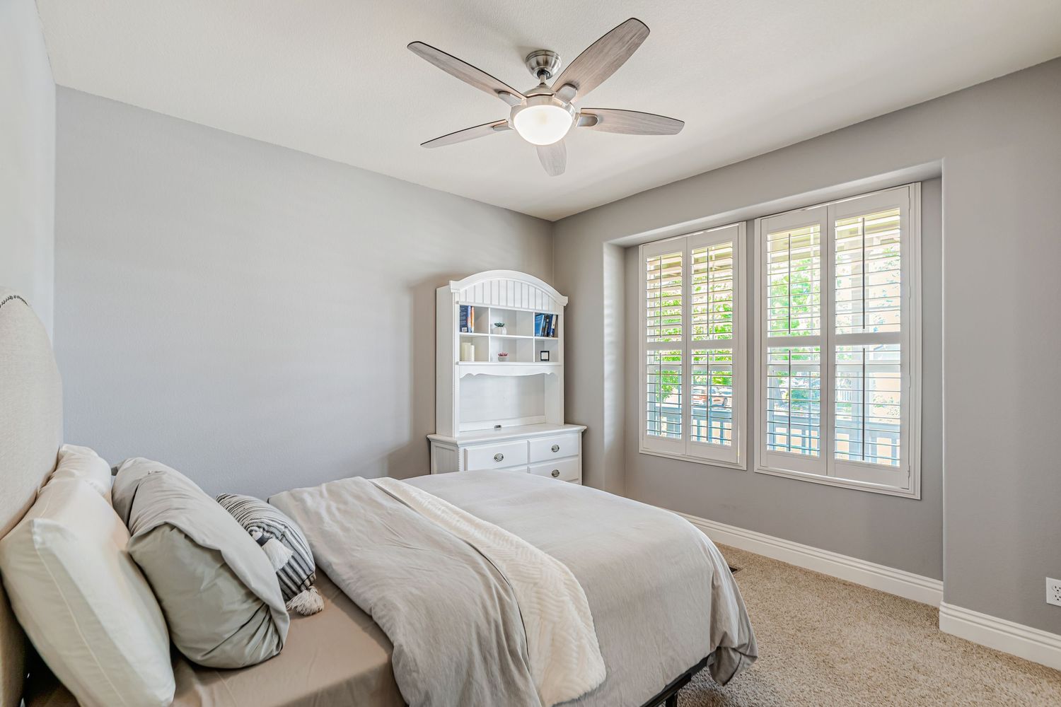 Bright bedroom with white built-in bookshelf, plantation shutters, and ceiling fan.