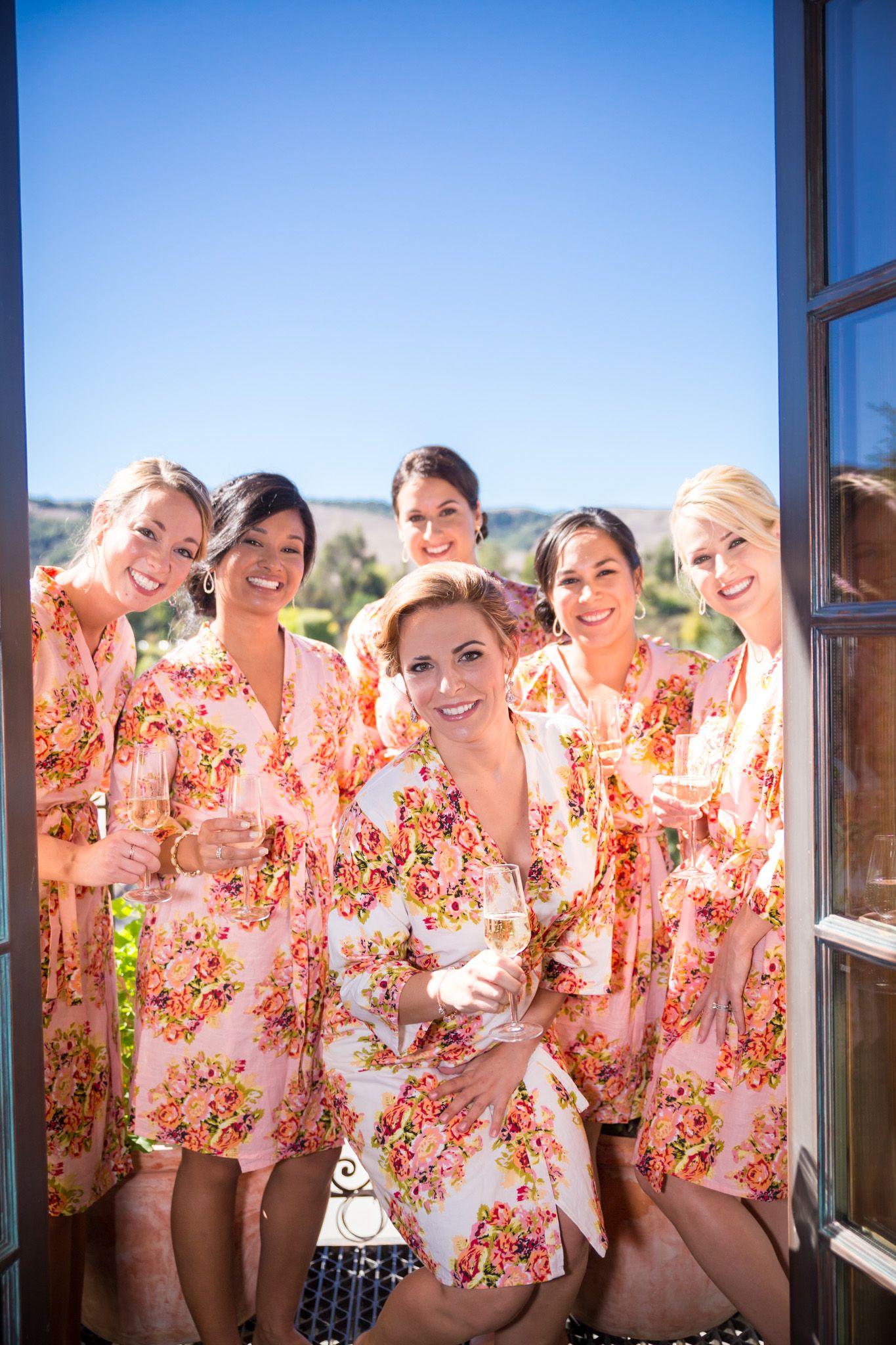 Bridesmaids wearing matching floral pink robes pose together by a window with mountain views.