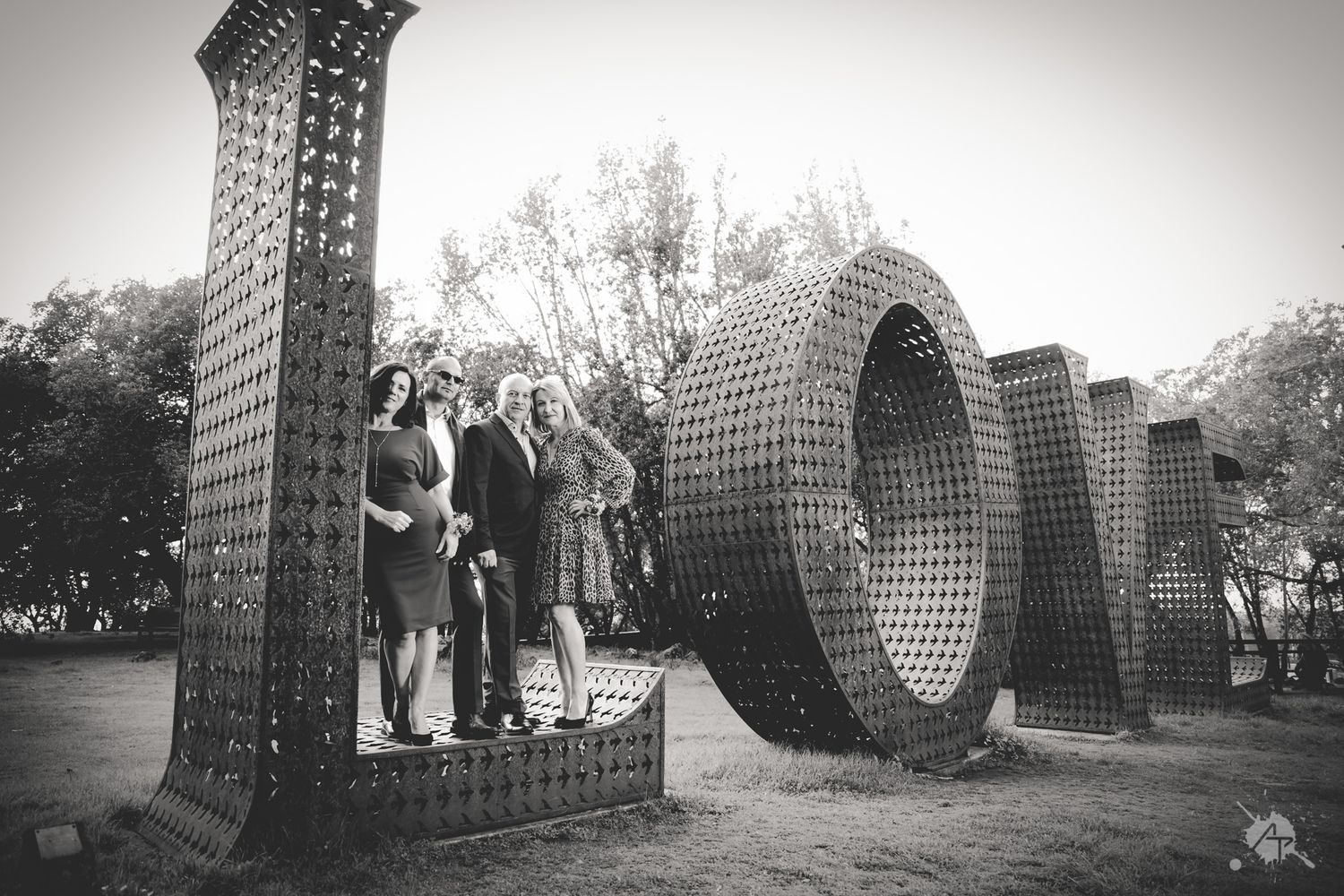 Black and white photo of people posing with large sculptural LOVE letters made from an intricate woven pattern outdoors.