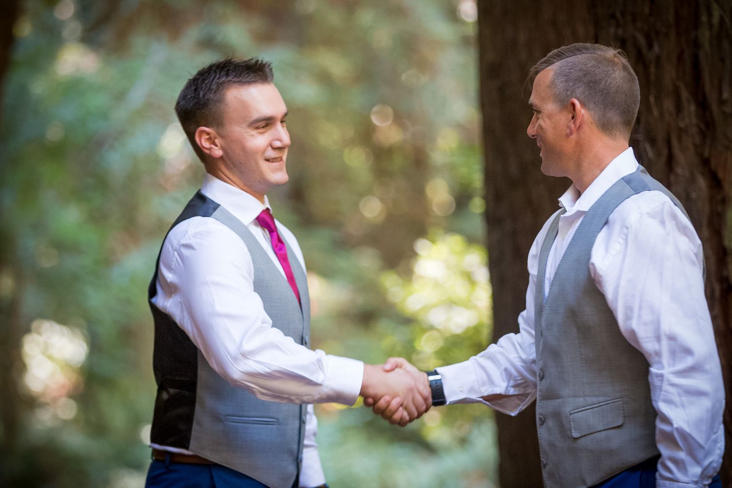 Two groomsmen in grey vests shake hands in wooded outdoor setting.