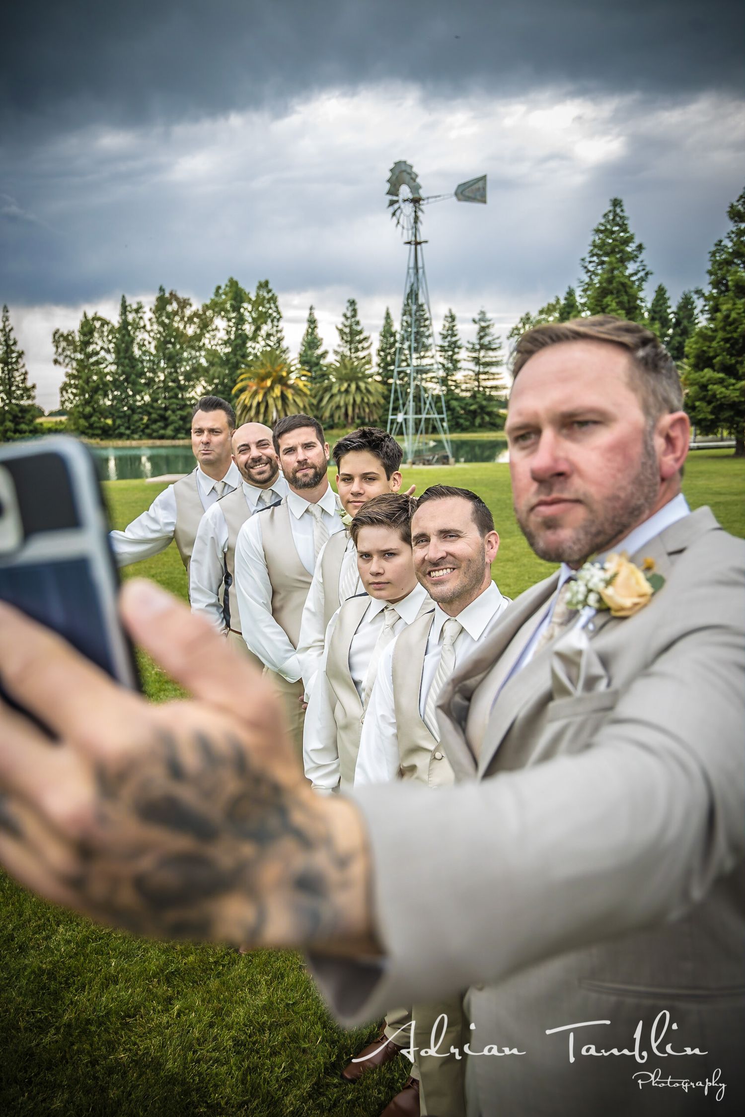A wedding party takes a group selfie outdoors with a windmill and stormy sky in the background.