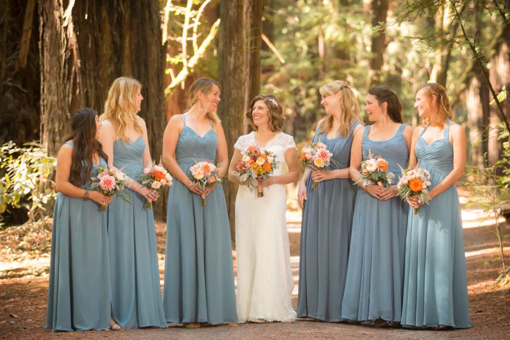 Bridesmaids in matching blue gowns pose with bride holding coral and peach bouquets in forest setting.