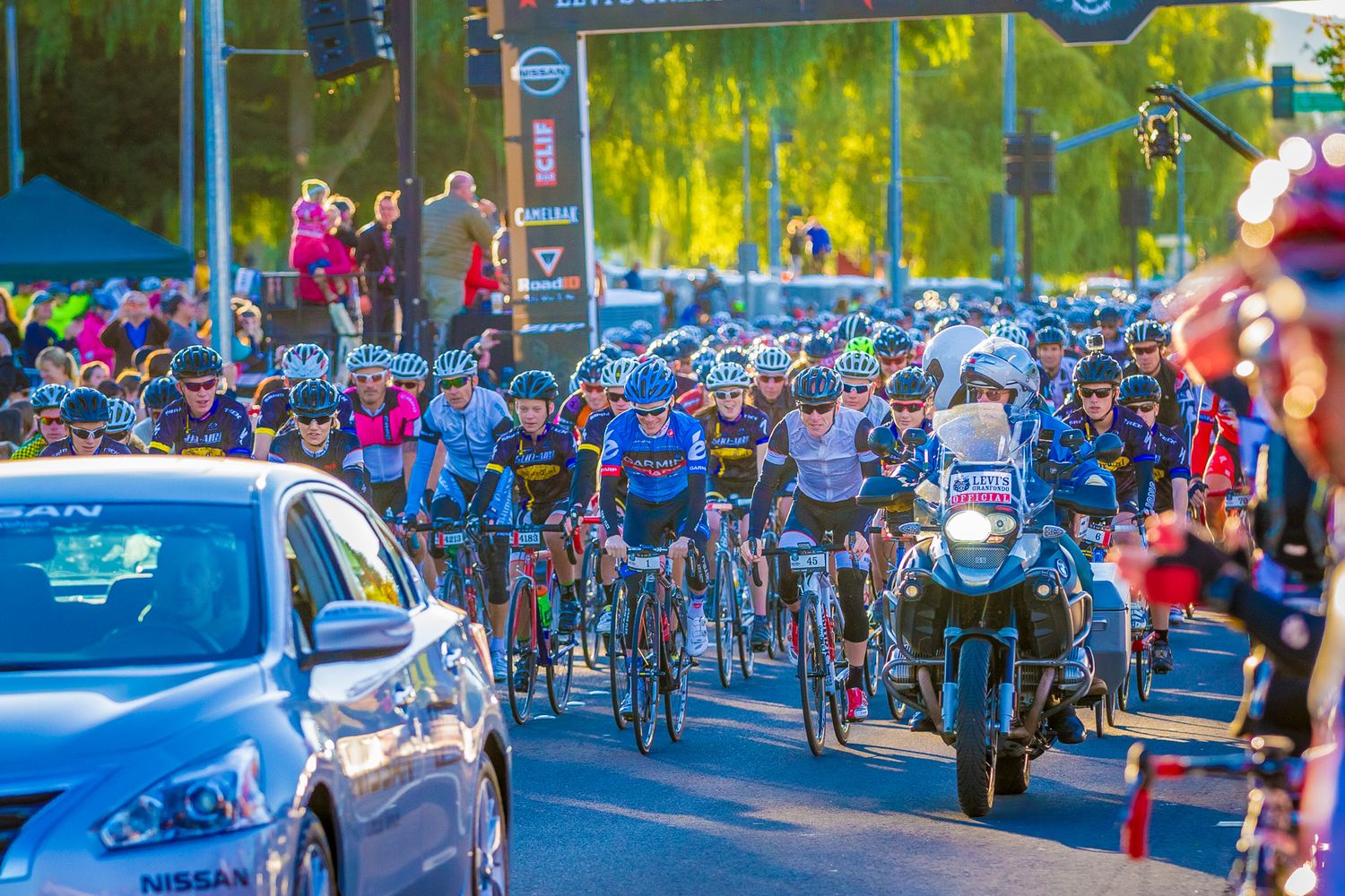 Large crowd of cyclists gathered at a street event with cars and motorcycles visible.