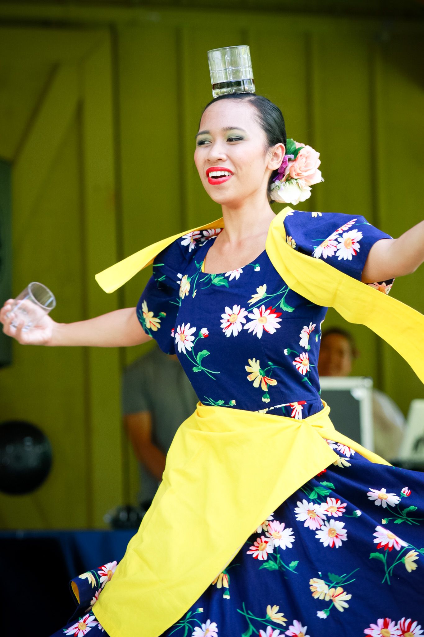 Dancer in navy floral dress with yellow apron performs traditional dance with a bottle balanced on her head.
