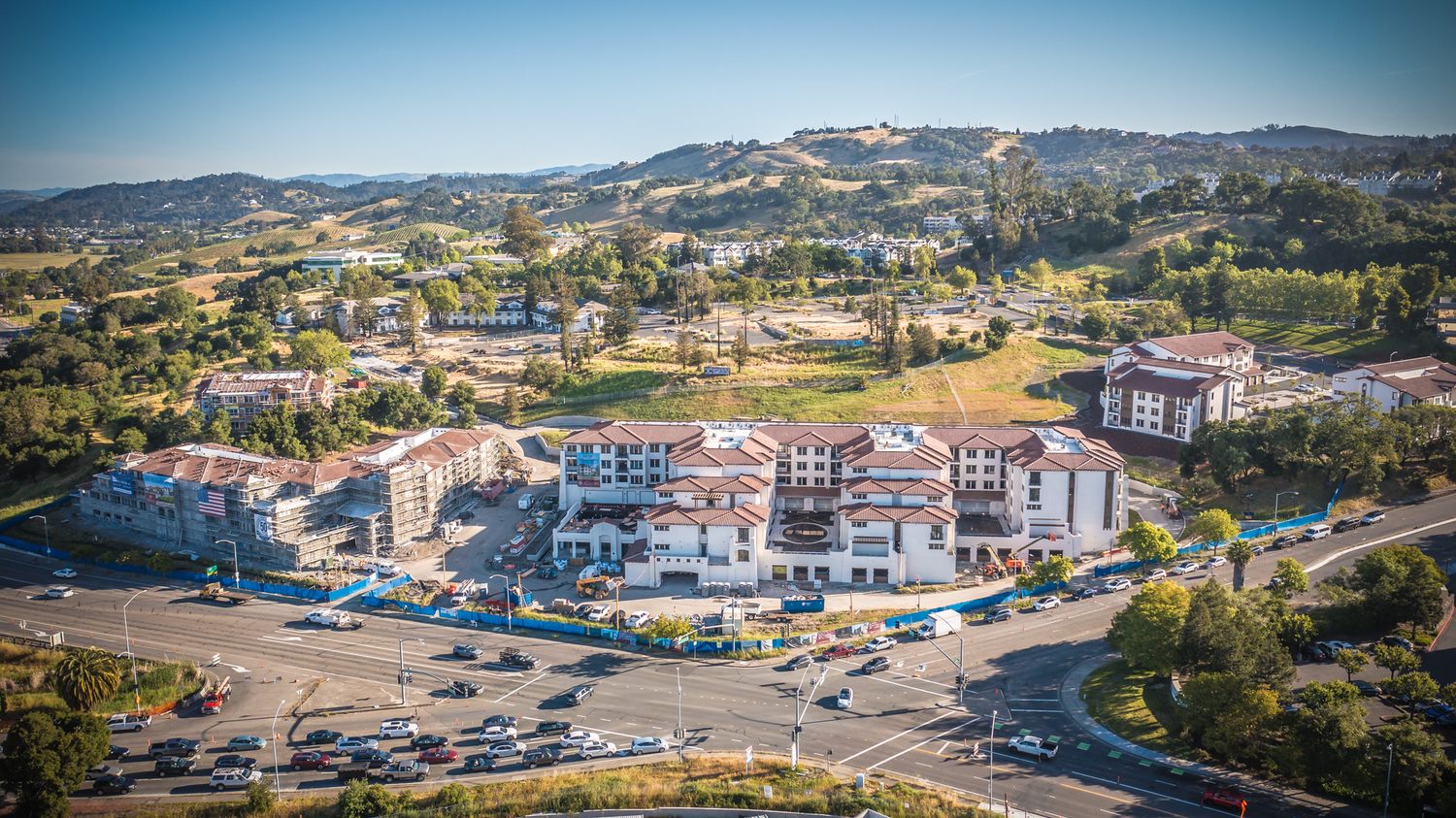 Mediterranean-style shopping center and parking area nestled in rolling hills with mountains in distance.