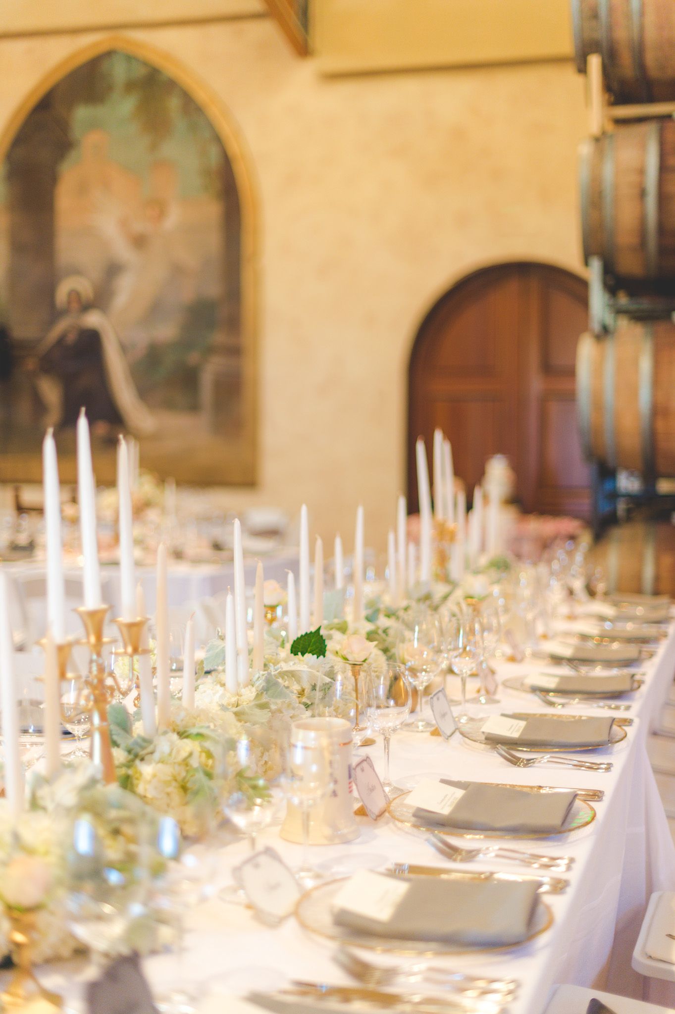 An elegant wedding reception table set with white candles, floral centerpieces and place settings in a wine cellar room.