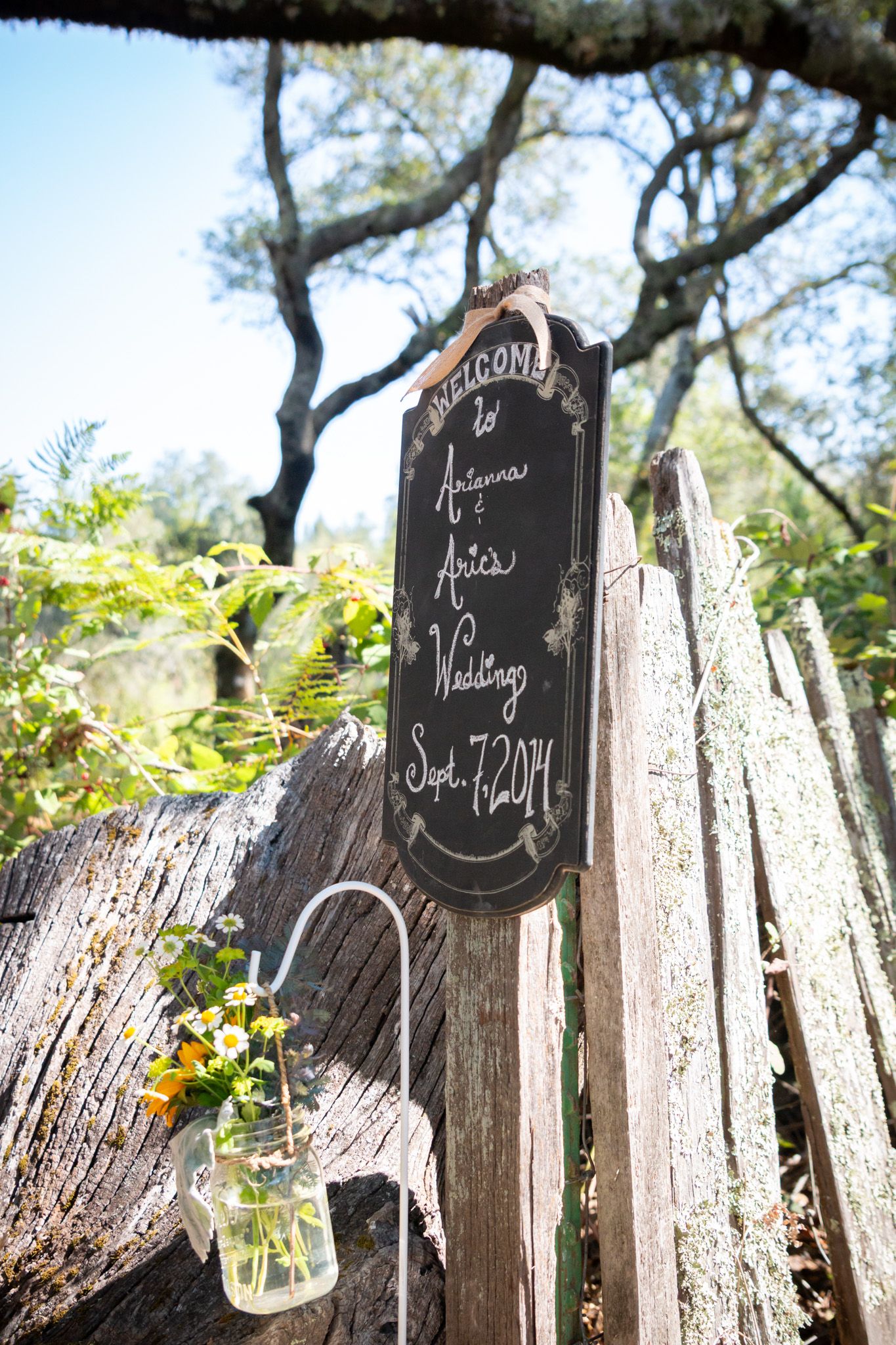 Rustic welcome sign and mason jar flower vase hang on wooden fence for outdoor wedding decoration.