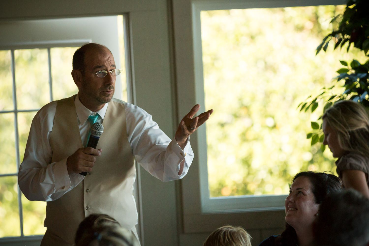 Person gestures while speaking at a business meeting with others gathered around a sunlit conference table.