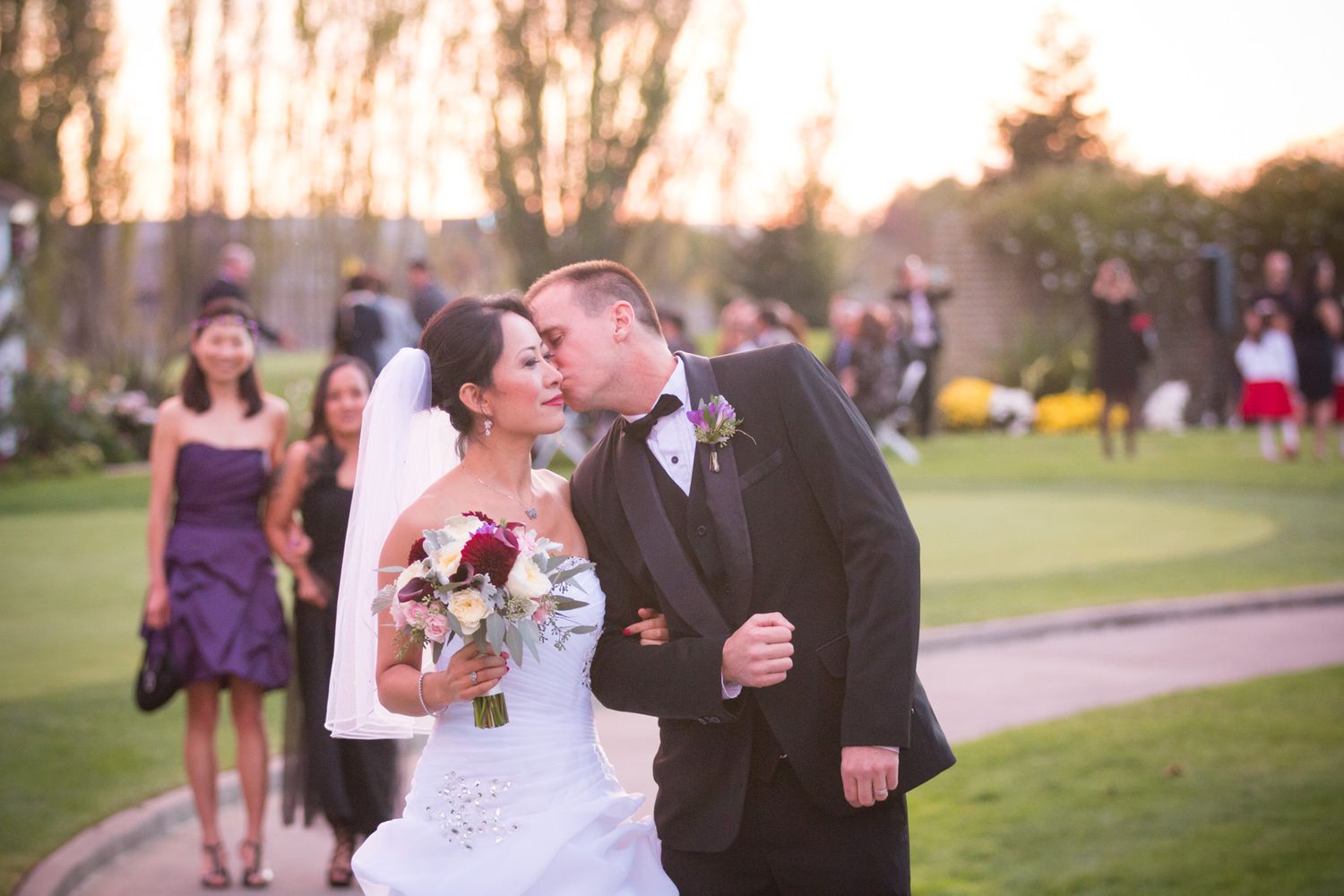 A romantic outdoor wedding moment captured on a golf course at sunset with guests in the background.