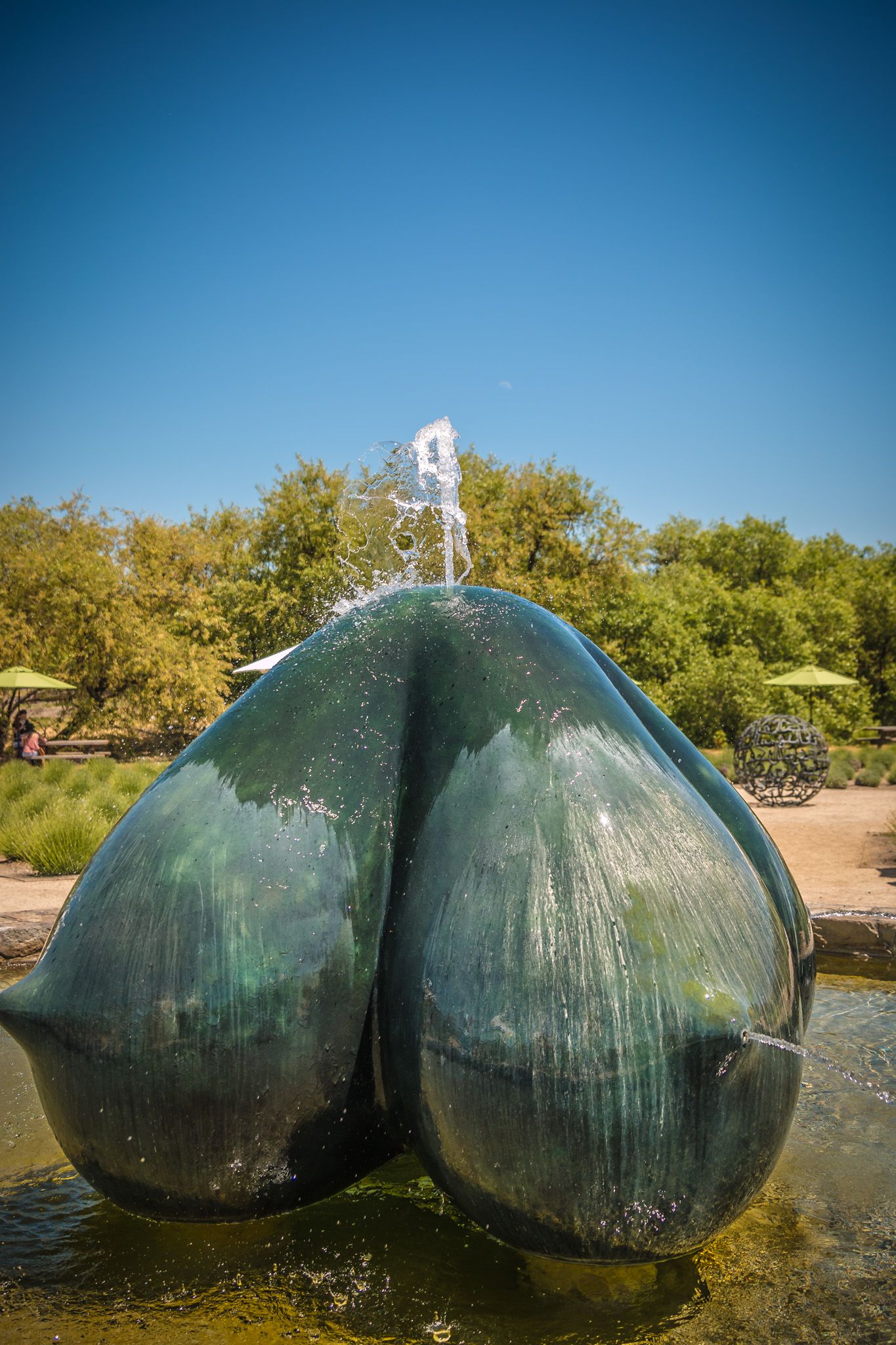 Large green sculptural fountain with water spraying from the center in a garden setting.