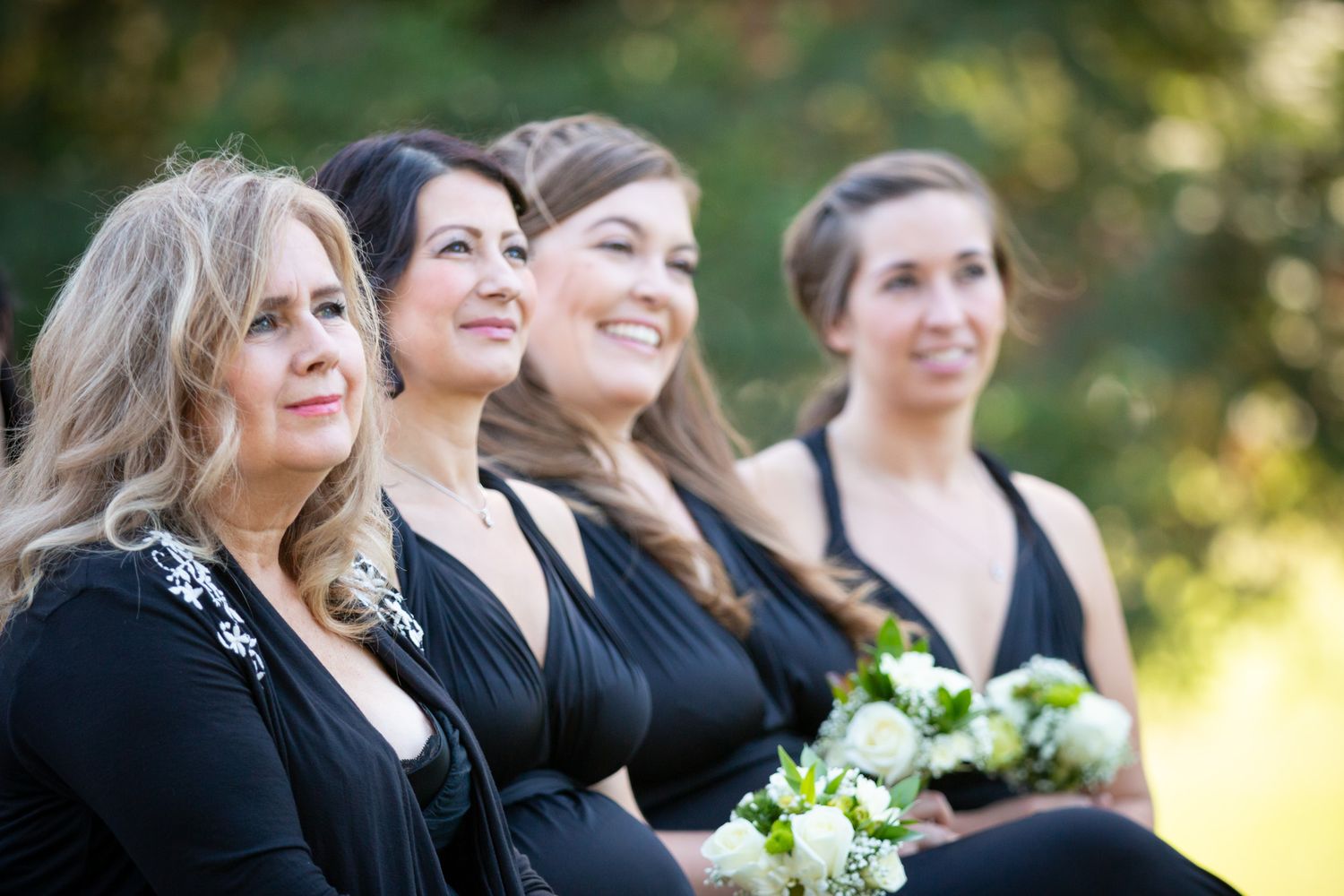 Bridesmaids in black dresses smile while holding white floral bouquets.