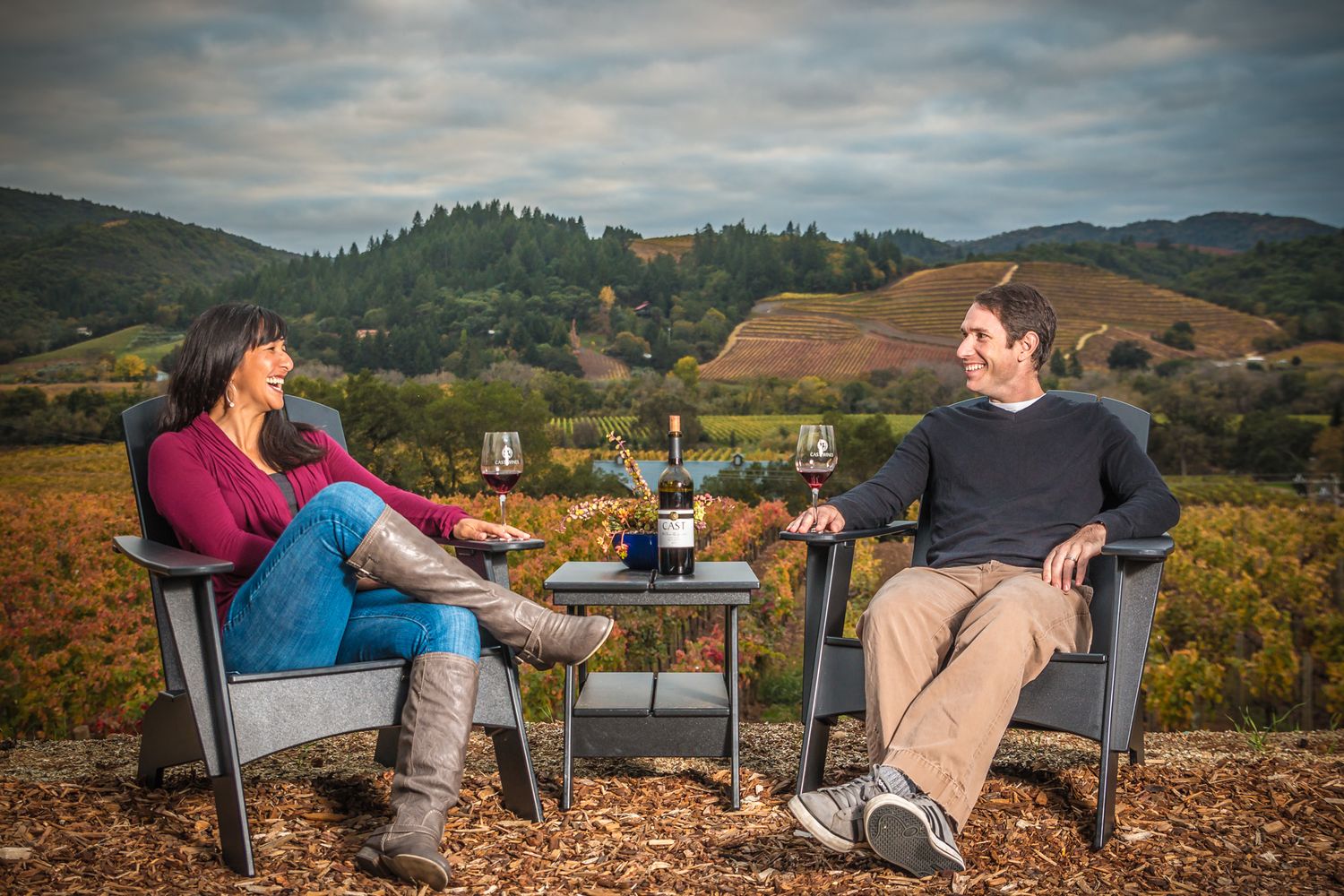 Two people enjoying wine and conversation on outdoor chairs overlooking vineyard hills at sunset.