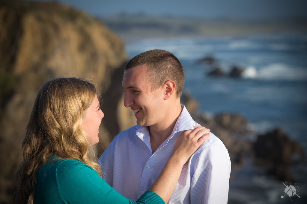 A couple embraces along a rocky coastline with crashing waves and cliffs in the background during golden hour.