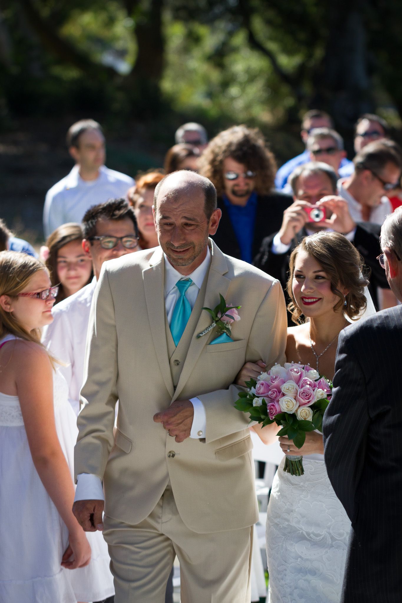 Groom in tan suit and blue tie walks with pink bouquet during outdoor garden ceremony.