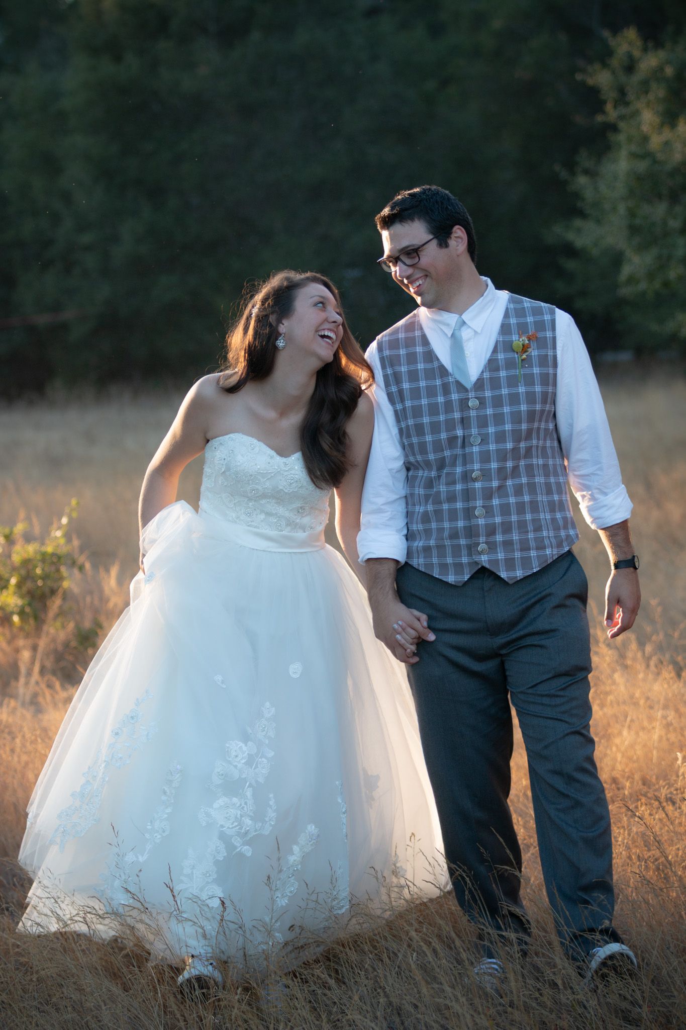 A couple walking together in a golden field at sunset, wearing wedding attire including a white dress and gray vest.