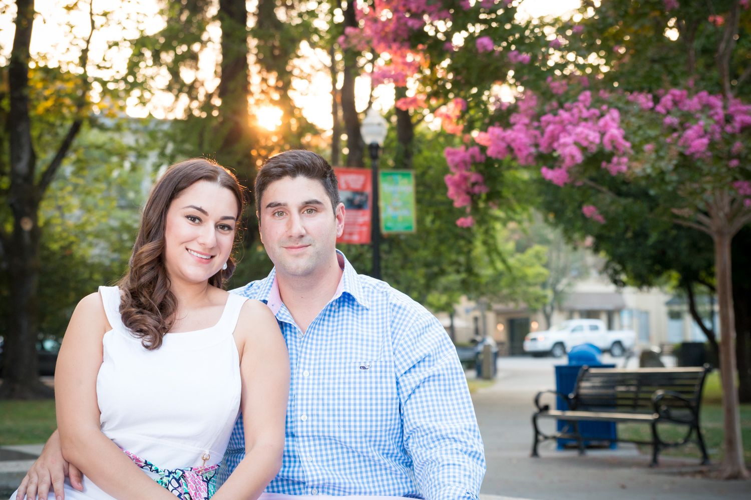 A couple sits together outdoors at sunset with pink flowering trees and benches in a scenic park setting.