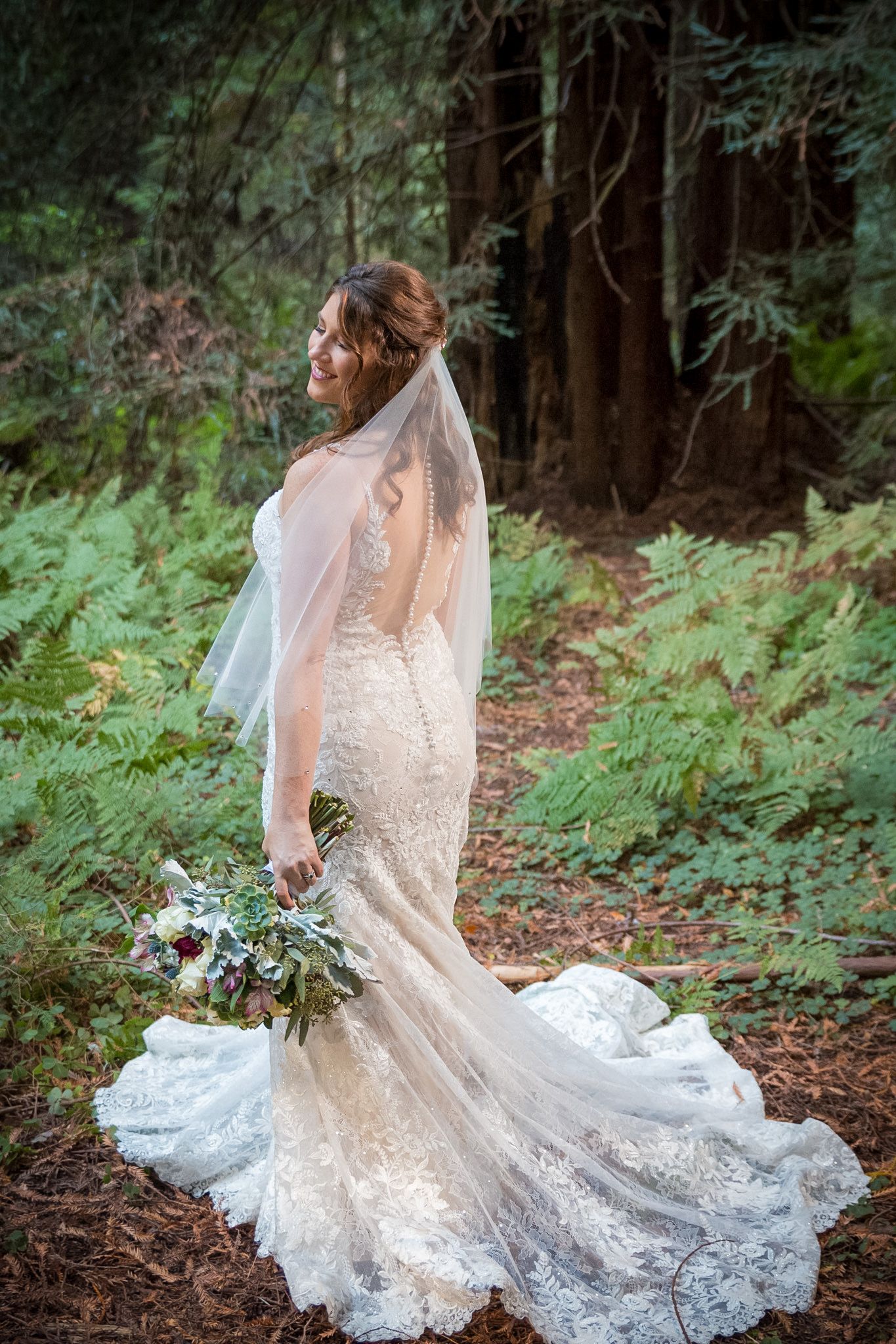 Bride in lace wedding gown with long train poses in enchanted forest setting.