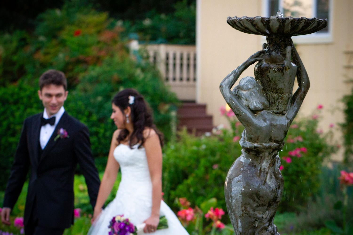 Garden statue in foreground with wedding couple posing among blooming flowers.