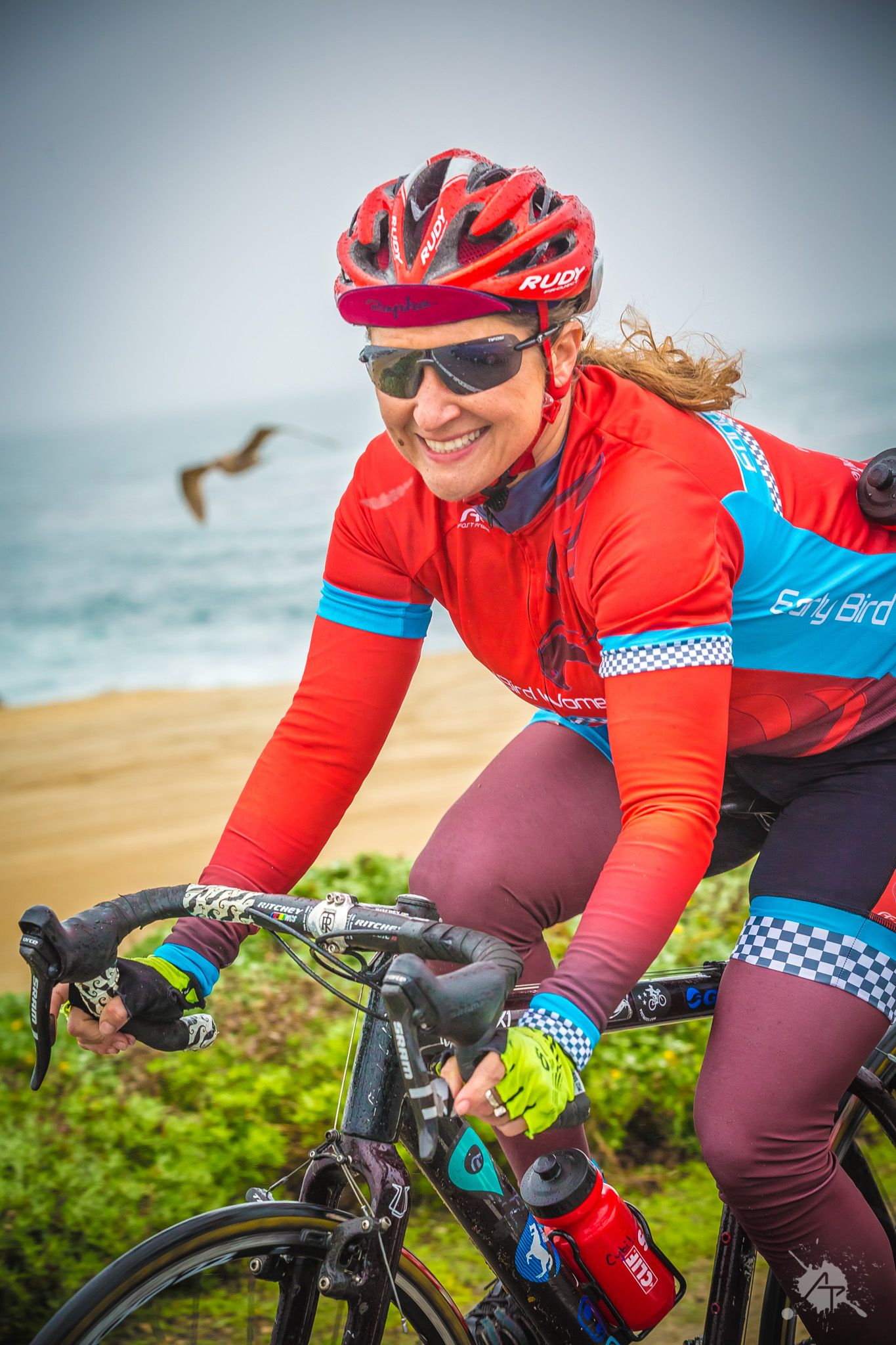 Road cyclist wearing red helmet and sunglasses rides along coastal trail with ocean and beach in background.