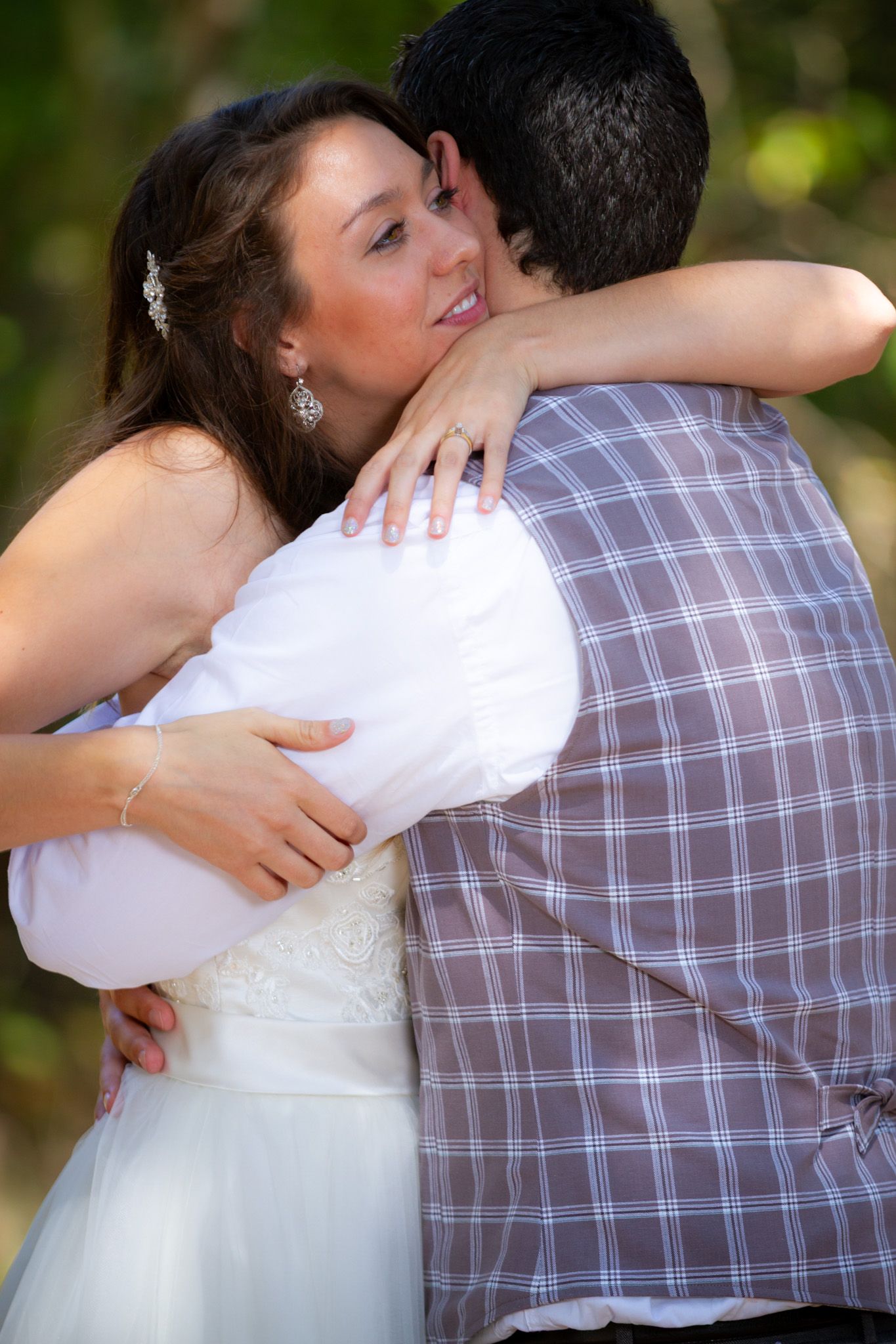A couple embraces during what appears to be a wedding photoshoot, with the bride wearing a white dress and groom in plaid shirt.