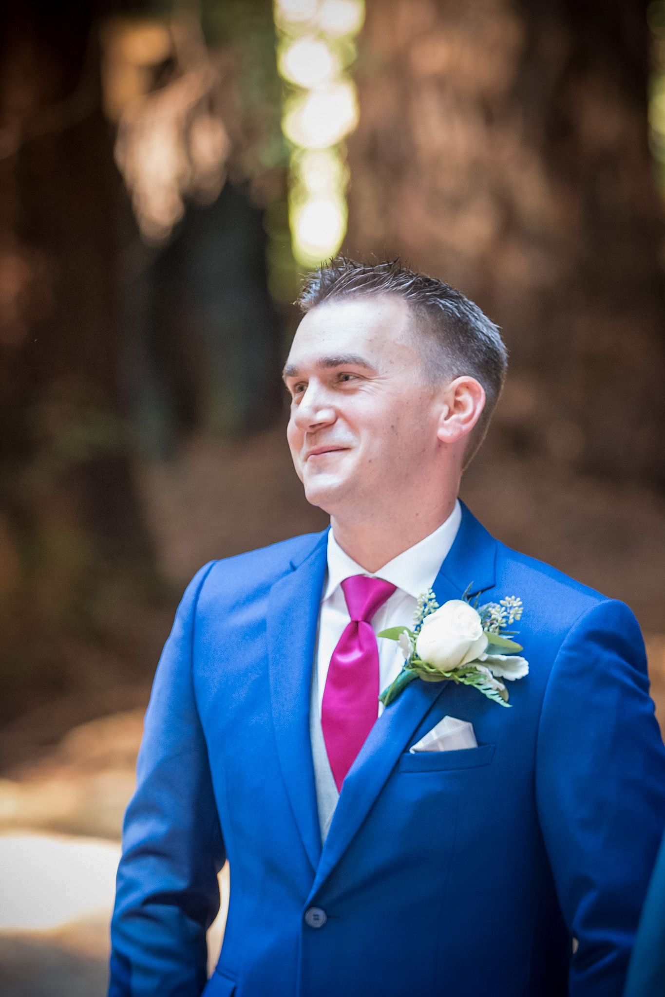 Groom in bright blue suit with pink tie and white boutonniere smiling outdoors.