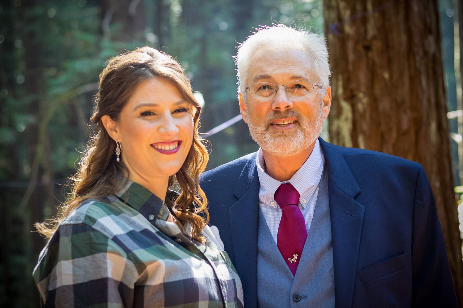 Two people pose together in a forest setting wearing contrasting casual and formal attire.
